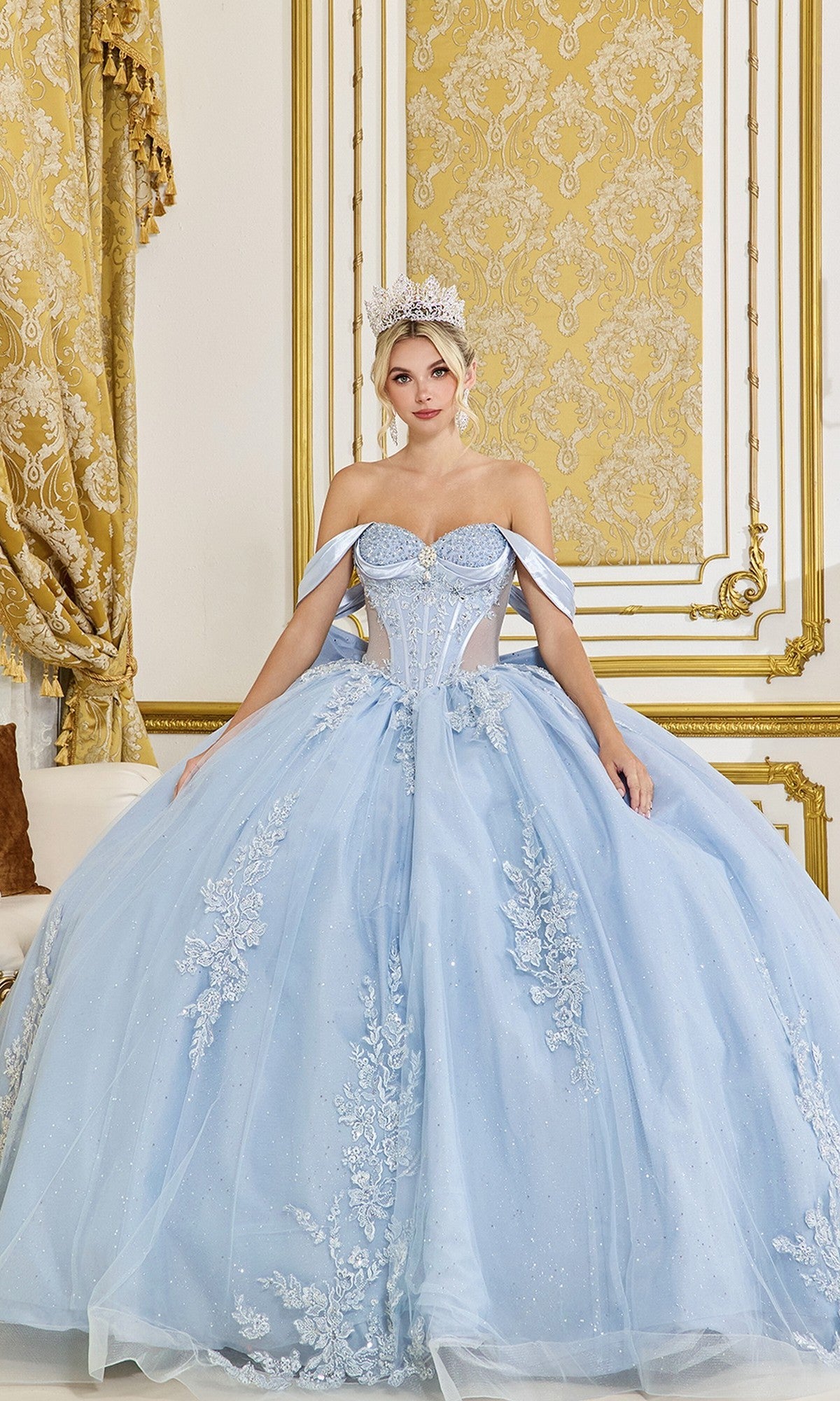 A woman in the Quinceanera Dress 5094 By Dancing Queen, a dusty blue gown with a corset bodice and tiara, stands in an ornate room adorned with gold accents and patterned wallpaper.